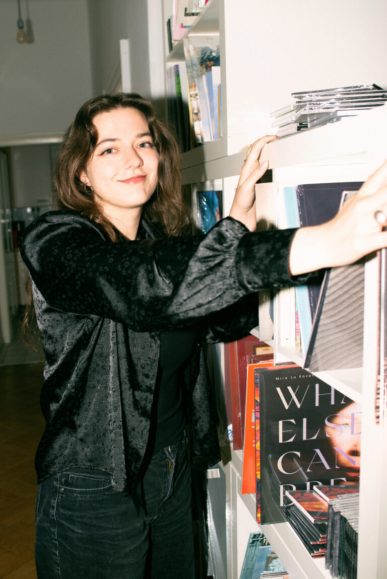 Ink Music - Teamfoto - Lily Strasser - steht vor einem Regal voll mit Cds, Bücher und LPs und schaut direkt in die Kamera