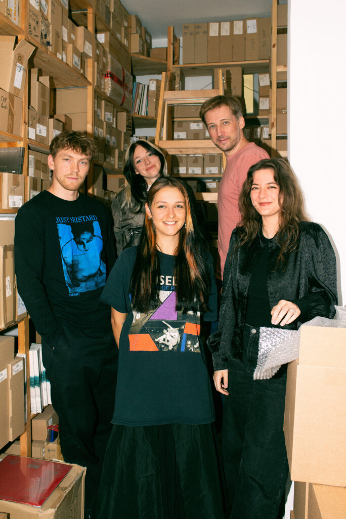 Ink Music - Teamfoto - 5 Menschen stehen in einem Lagerraum voll mit Kisten und blicken direkt in die Kamera