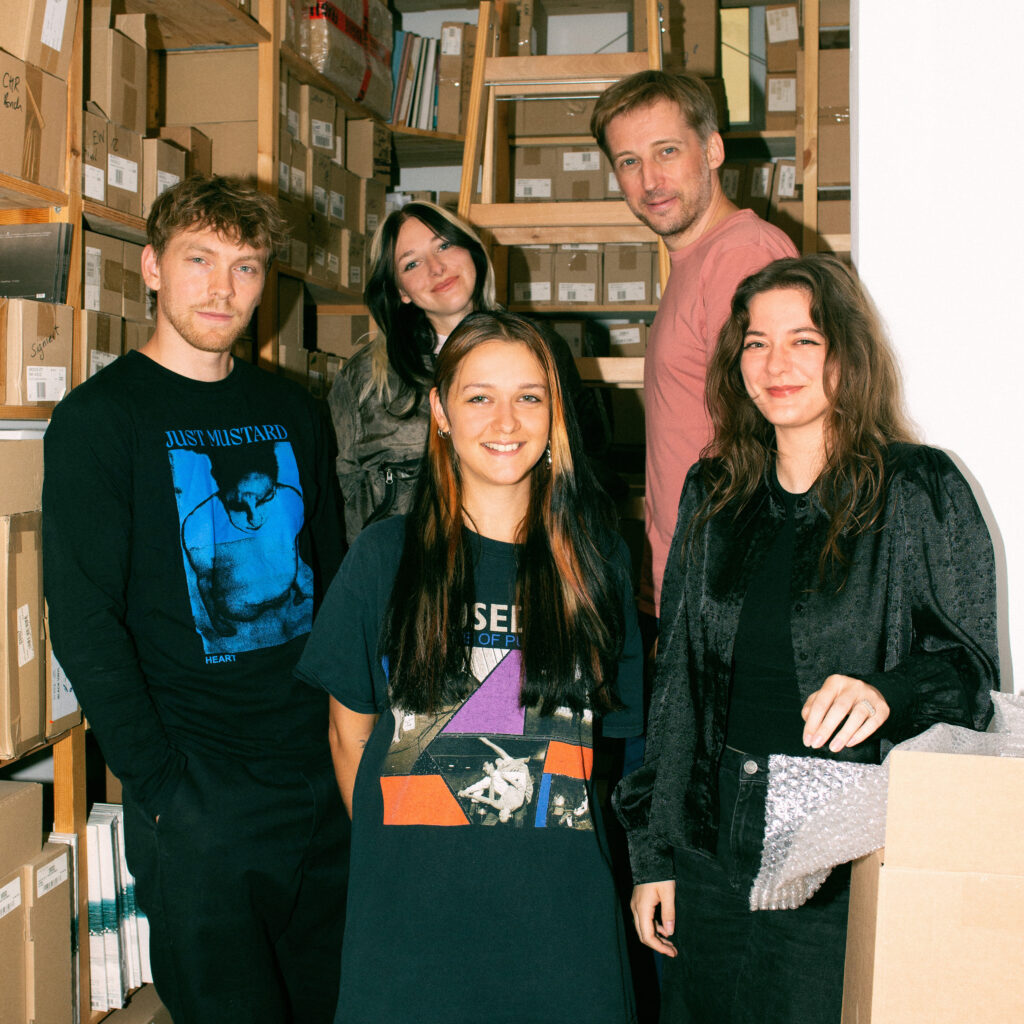 Ink Music - Teamfoto - 5 Menschen stehen in einem Lagerraum voll mit Kisten und blicken direkt in die Kamera